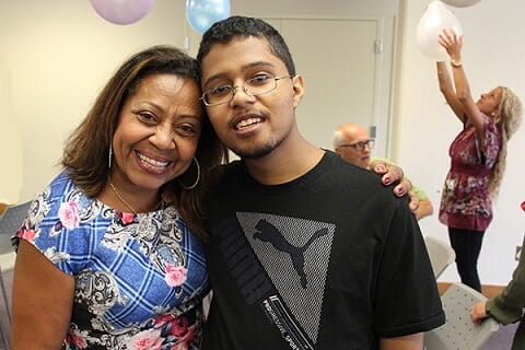 A man of colour and a woman of colour standing next to each other and smiling with balloons in the background. 