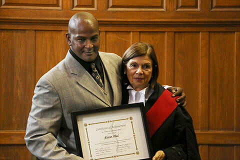 A man of colour in a suit and a Caucasian woman with short hair in a gown, both holding the graduation plaque smiling at the camera. 