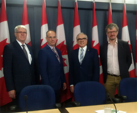 Four men in suits standing in front of four Canadian flags.