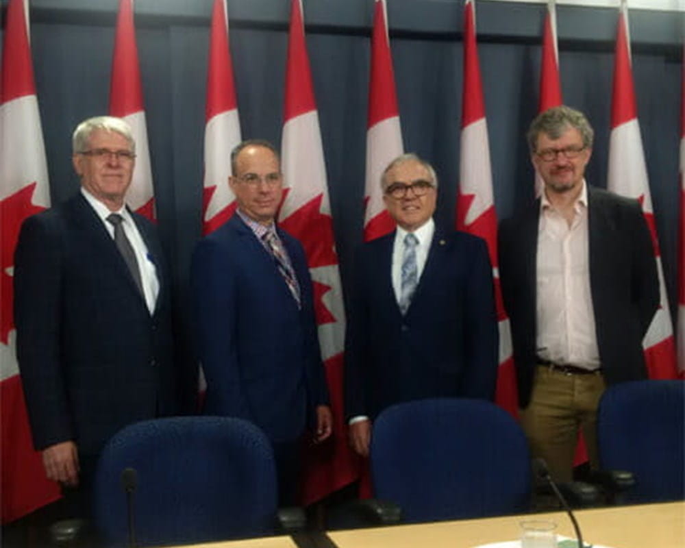 Four men in suits standing in front of four Canadian flags.