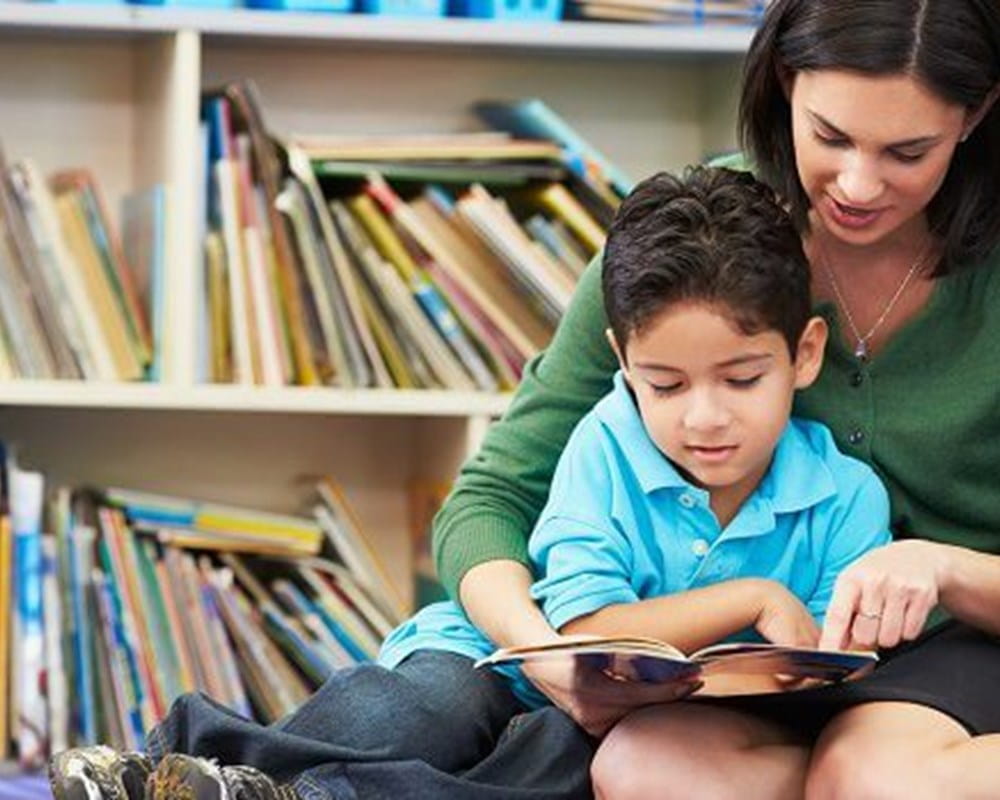 A child and a woman sitting down and reading a book together in a library.