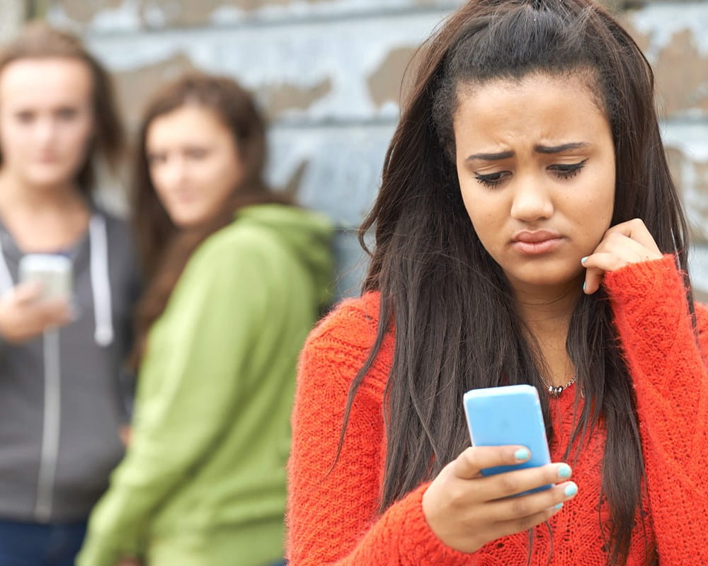 Girls looks sadly at her phone with other girls gossiping in background