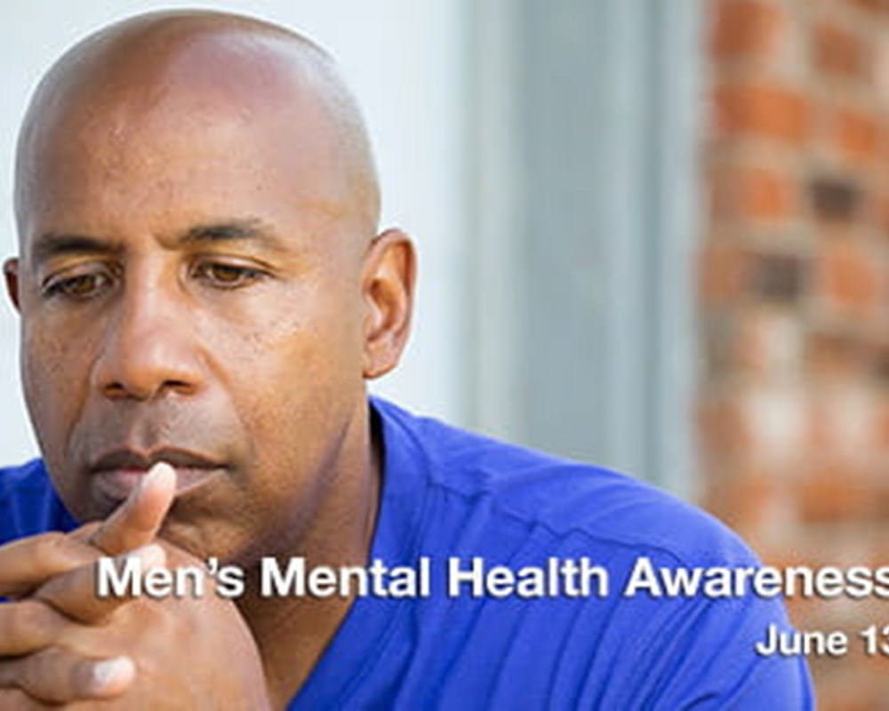 A bald man of colour wearing a blue t-shirt clasping his hands in front of his face looking upset.