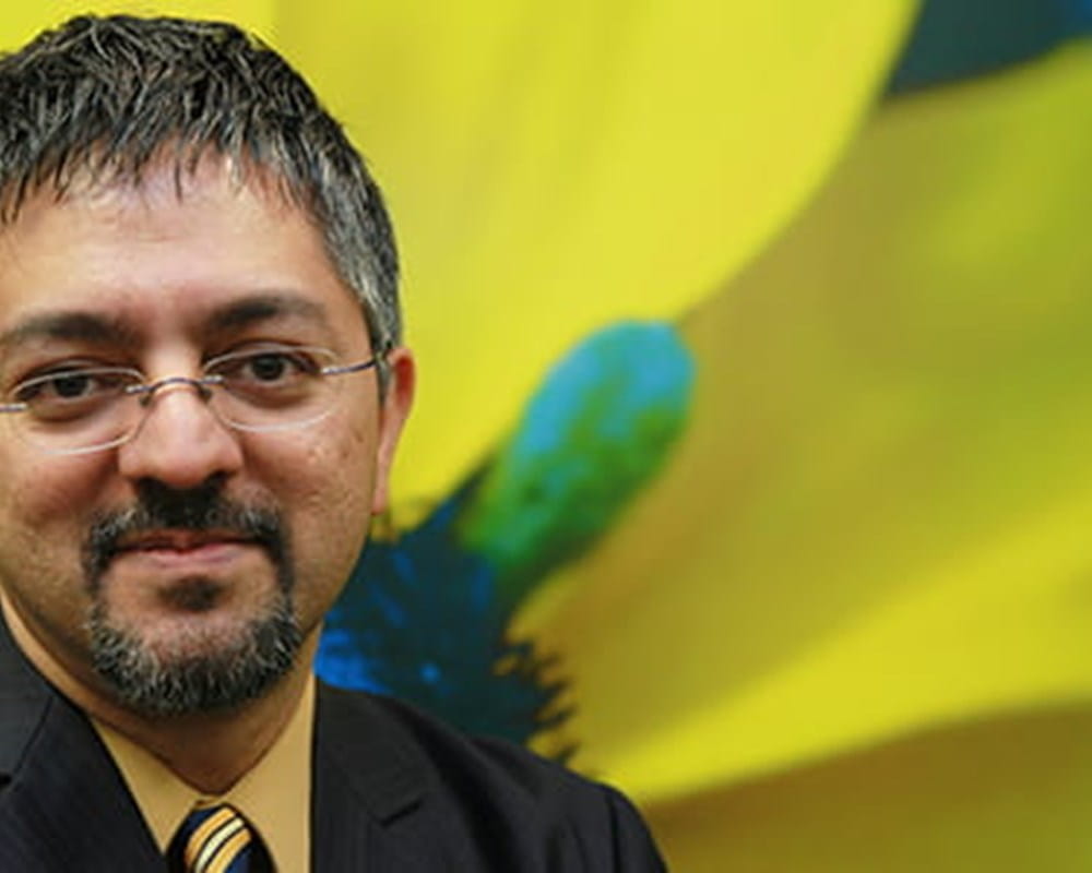 A coloured man with glasses smiling in front of an image of a flower with yellow petals.