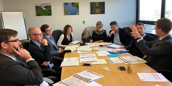Eight men and women in business clothes sitting in a boardroom having a meeting with binders and papers all over the table. 