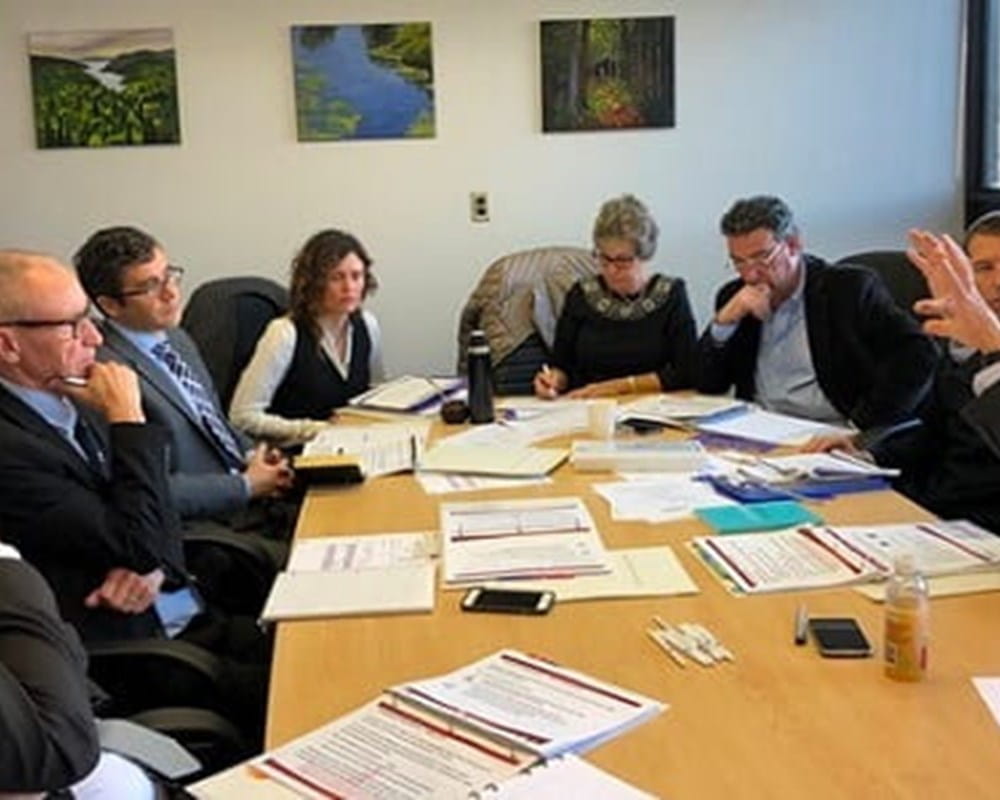 Eight men and women in business clothes sitting in a boardroom having a meeting with binders and papers all over the table.