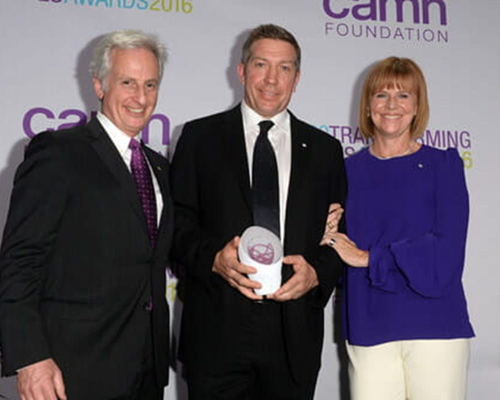 Two men in business suits and a woman in a blue blouse standing beside each other smiling while holding a white award.