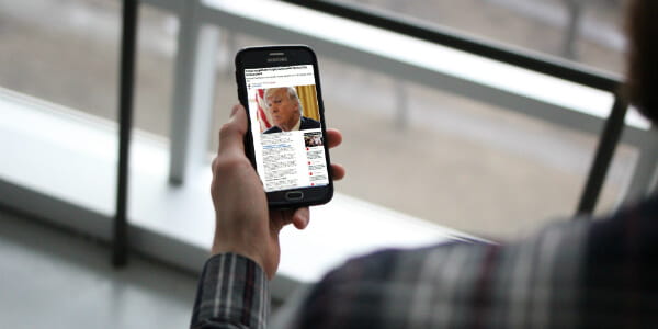 A man wearing a long sleeve plaid shirt holding a phone with a picture of Donald Trump in a news headline. 