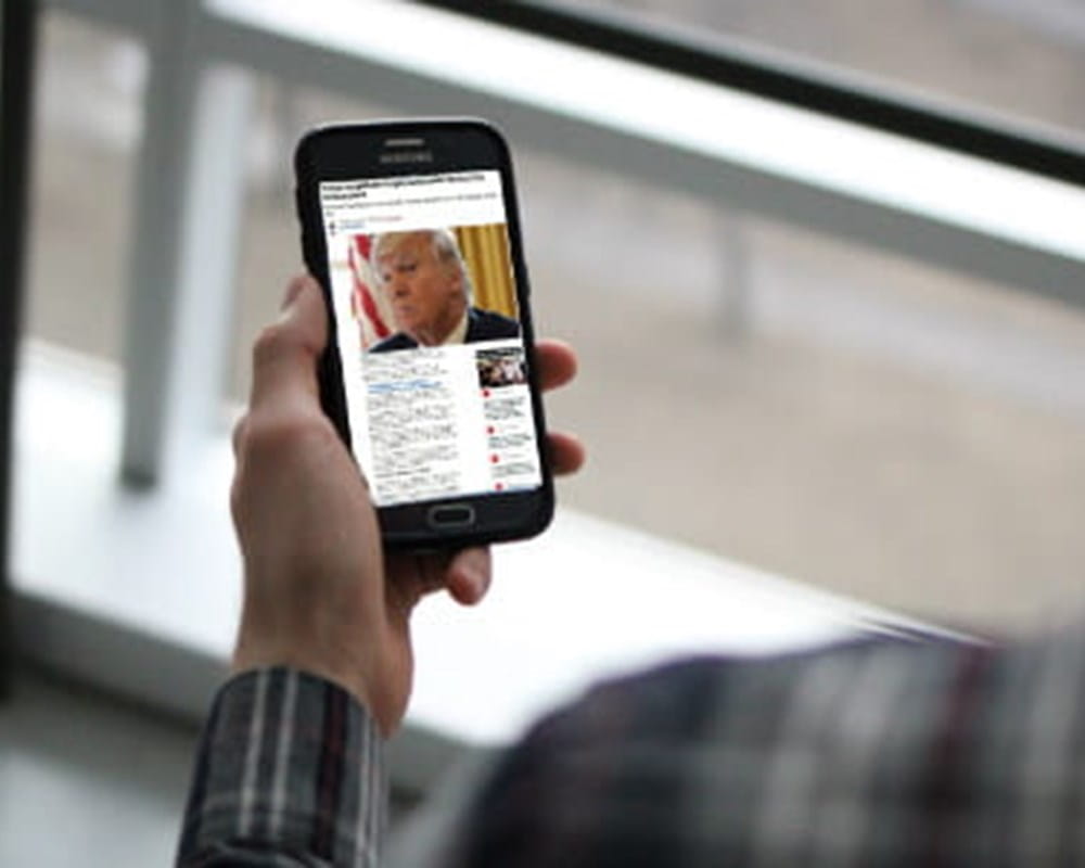 A man wearing a long sleeve plaid shirt holding a phone with a picture of Donald Trump in a news headline.