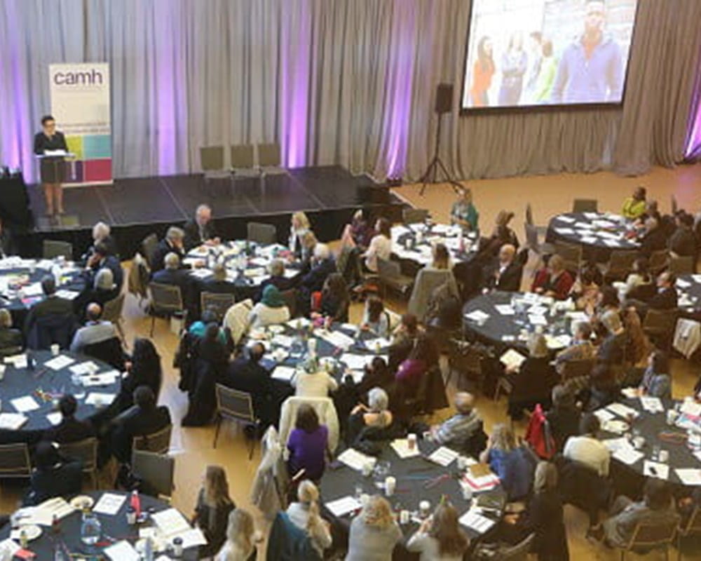 Several black round tables with people dressed in business clothes sitting around them, watching a person speaking in front of the podium on stage.